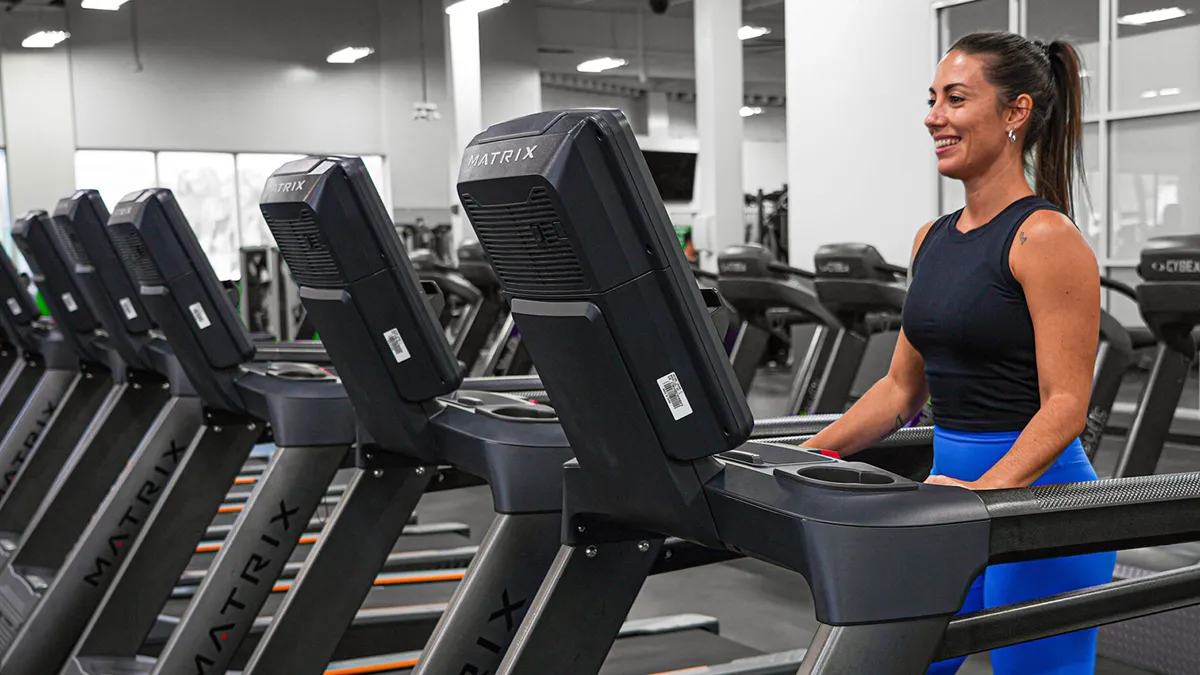 Smiling woman in blue leggings using a treadmill in a modern gym with multiple Matrix treadmills lined up.