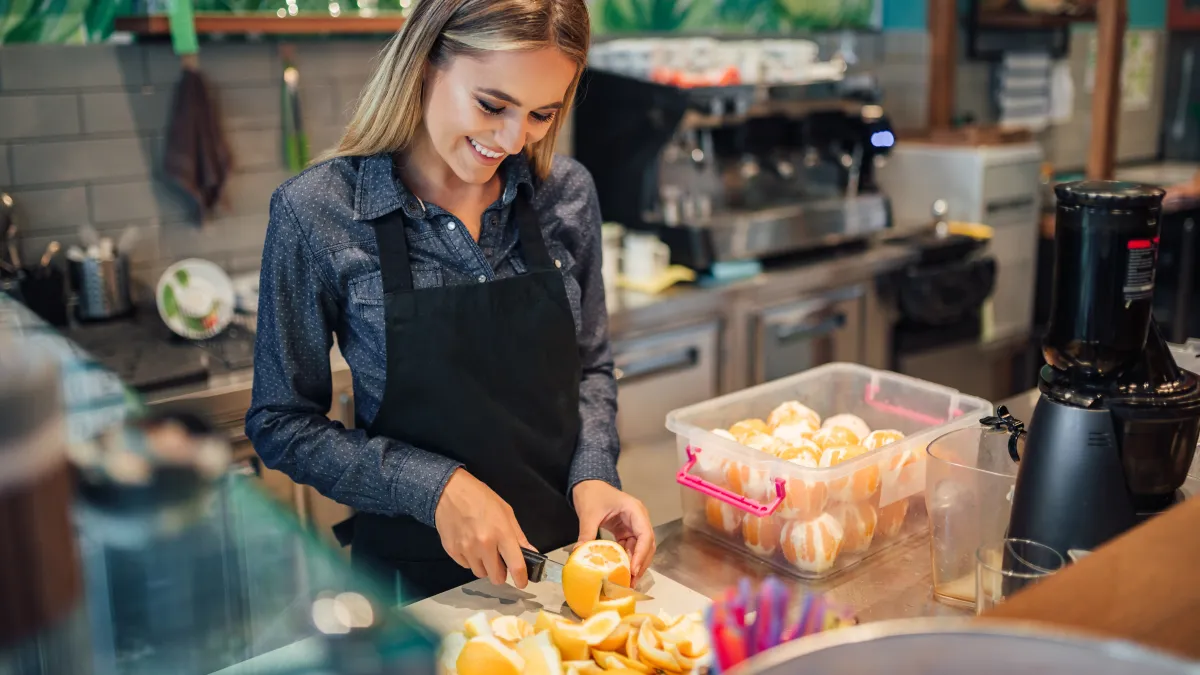 Smiling barista in black apron slicing fresh oranges behind cafe counter with juicer and juice container.