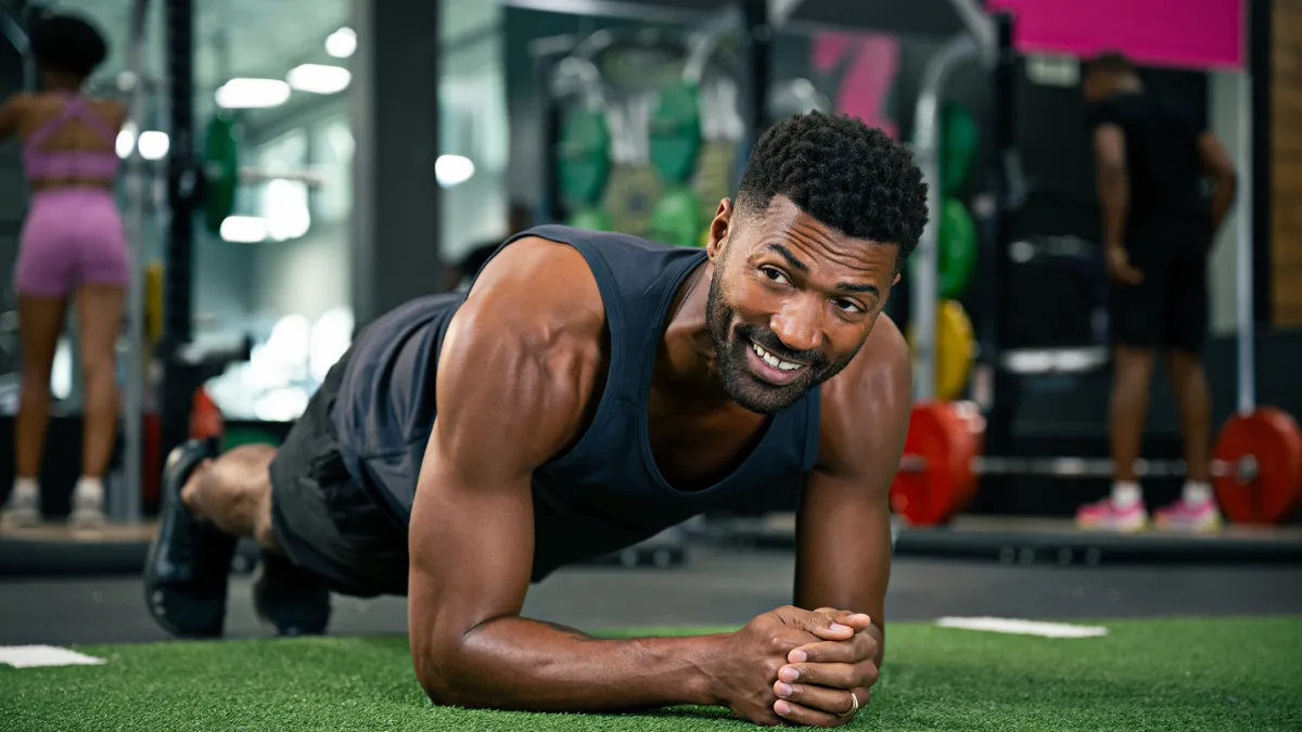 Fit man in black tank top doing plank exercise on green turf in gym with equipment in background