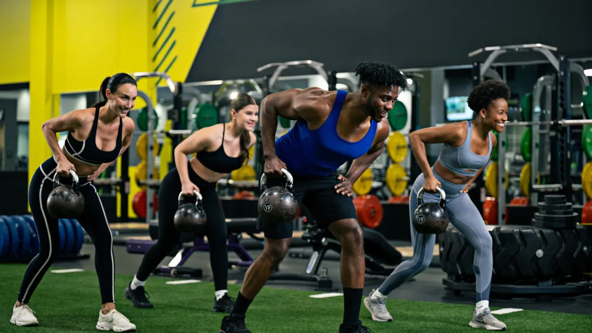 Four diverse adults exercising with kettlebells in a gym, performing bent-over rows on artificial turf.