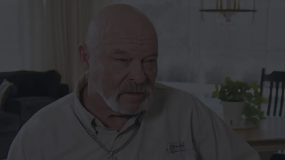 Elderly man with a white beard wearing a beige shirt and chain necklace, seated in a living room with plants and furniture.