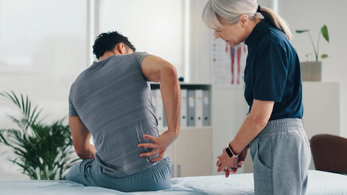 Male patient holding lower back in pain consulting elderly female doctor in a medical office clinic.