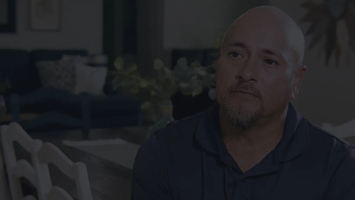 Man with a beard and mustache sitting thoughtfully at a dining table in a dimly lit room.