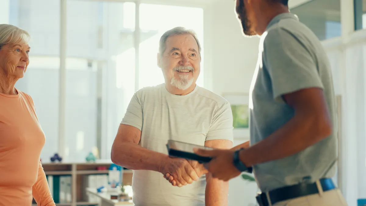 Older man smiling and shaking hands with a younger man while an older woman watches in a bright office