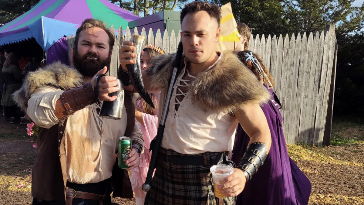 Two men in medieval attire holding drinking horns and cups, standing outdoors near tents and a wooden fence.