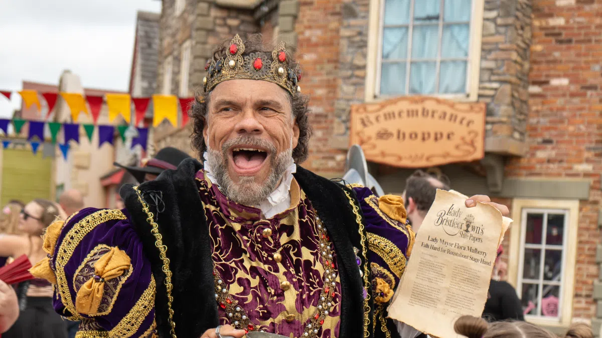 Man dressed as a medieval king with crown and robe holding a mug and a parchment at a Renaissance fair.