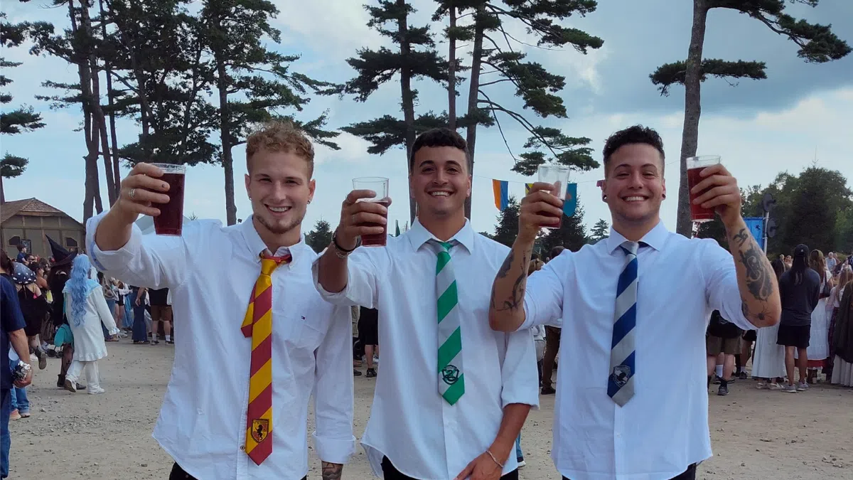 Three young men in white shirts and colorful ties holding drinks outdoors at a festive gathering with trees and crowd.