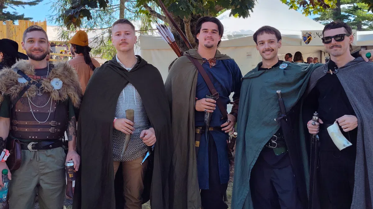 Five men dressed in medieval costumes with cloaks and weapons posing outdoors in daylight under trees.