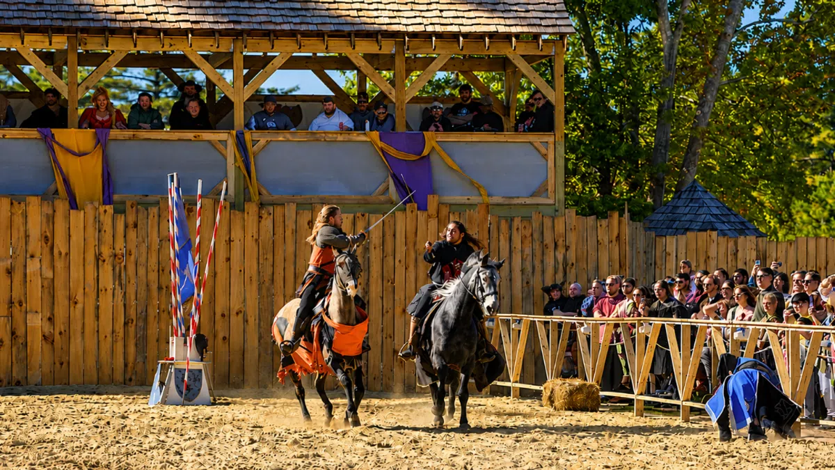 Two knights jousting on horseback in front of a wooden grandstand and a crowd on a sunny day