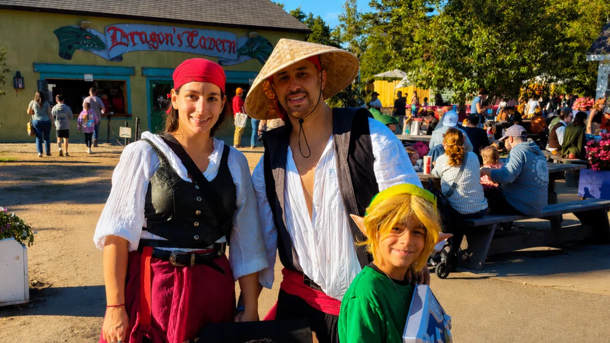 Family dressed in Renaissance and fantasy costumes posing outside Dragon's Tavern during a sunny festival day.