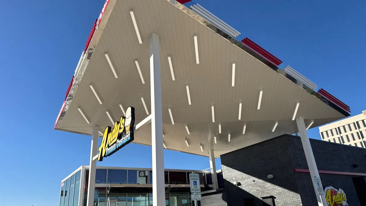 Low-angle view of a completed standing seam metal drive-thru canopy roof on an Andy's Frozen Custard restaurant in the Dallas-Fort Worth area, showing the finished underside with integrated lighting against a clear blue sky.