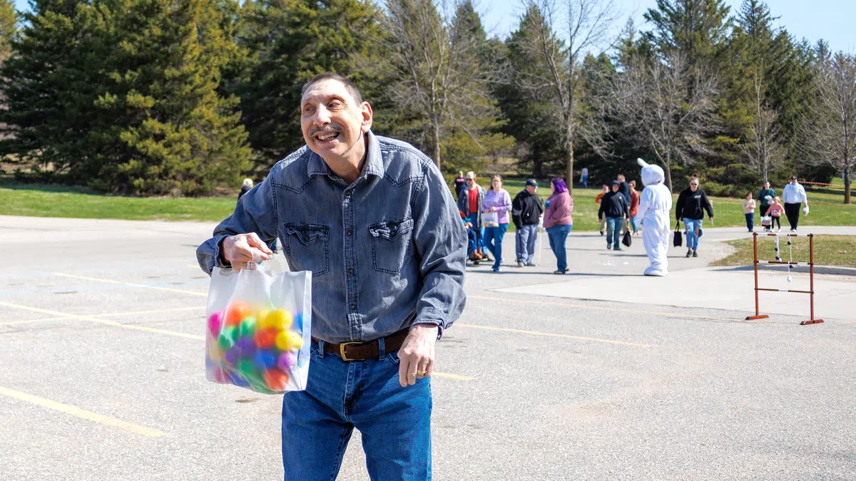 Smiling man holding a bag of colorful plastic eggs at an outdoor Easter egg hunt event on a sunny day.