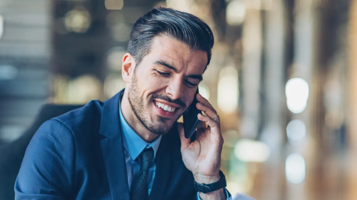 Smiling businessman in a suit talking on the phone while reviewing documents in a modern office setting.