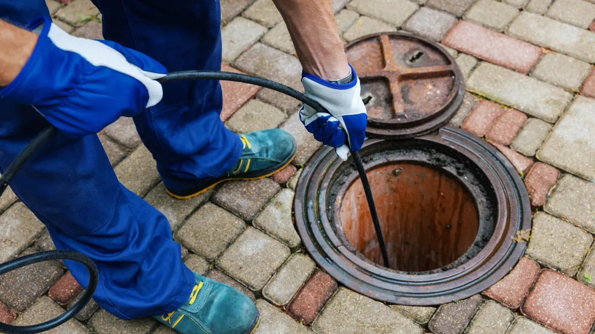 Worker in blue uniform and gloves clearing clogged drain using a plumbing snake on brick pavement.