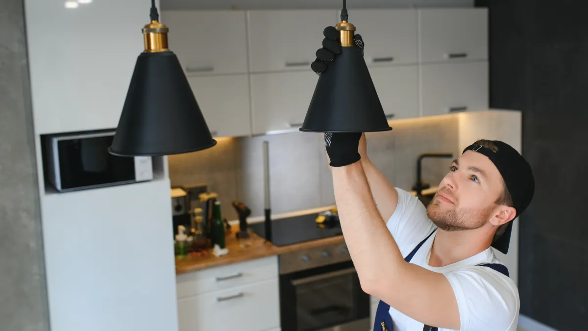 Man in work gloves installing black pendant light fixture in modern kitchen with white cabinets.