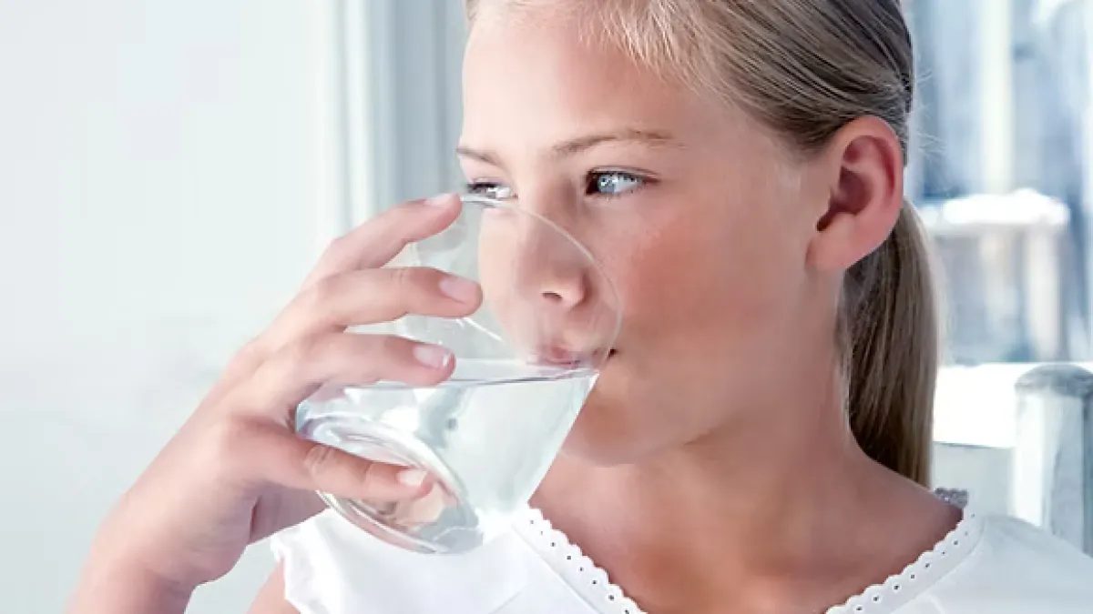 Young girl with blonde hair drinking water from a clear glass in a bright room.