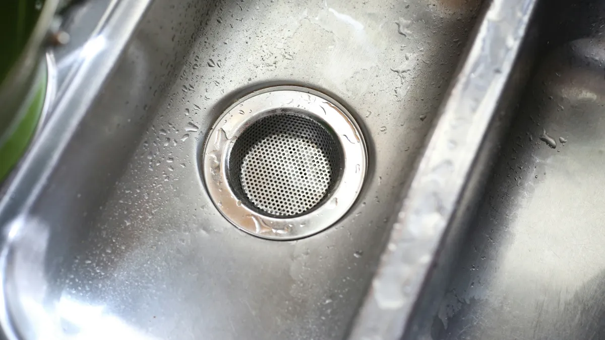 Close-up of a wet stainless steel kitchen sink drain with water droplets and reflections on the surface.