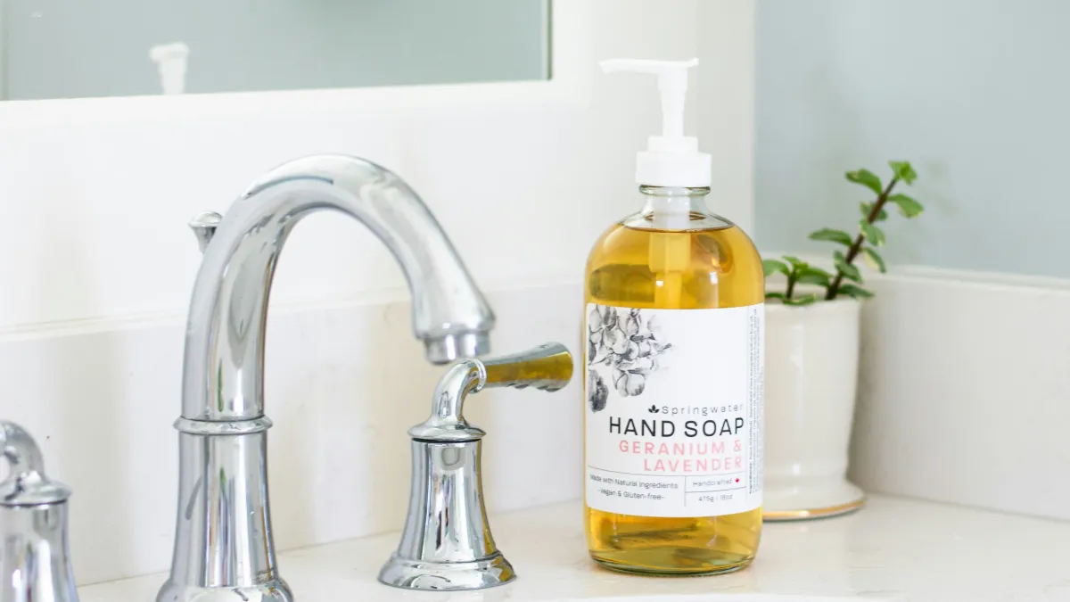 Clear glass bottle of geranium and lavender hand soap on white countertop beside chrome faucet and small potted plant