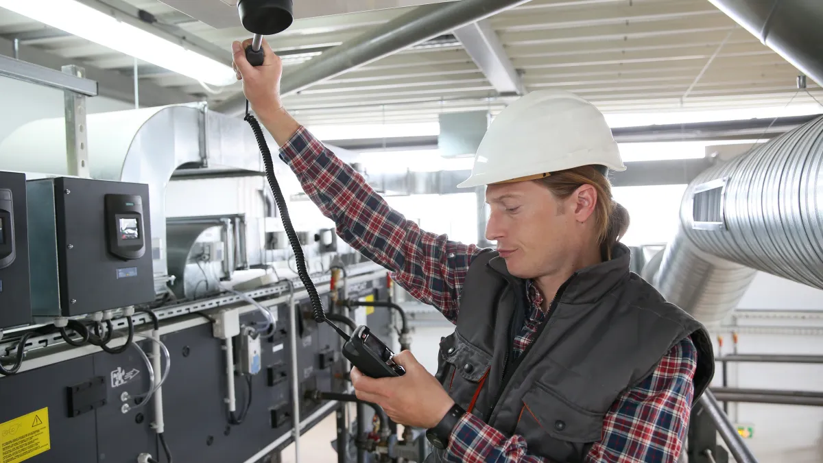 Technician in hard hat using device to inspect industrial HVAC system in mechanical room