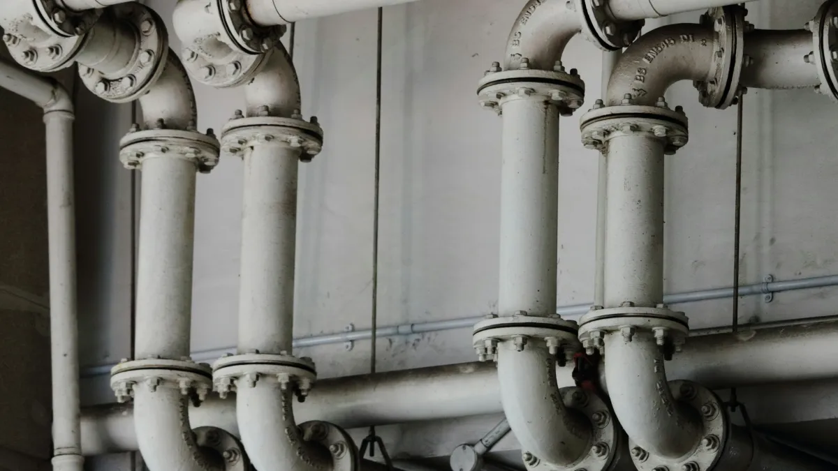 Industrial metal pipes with flange fittings arranged vertically and horizontally against a gray wall.