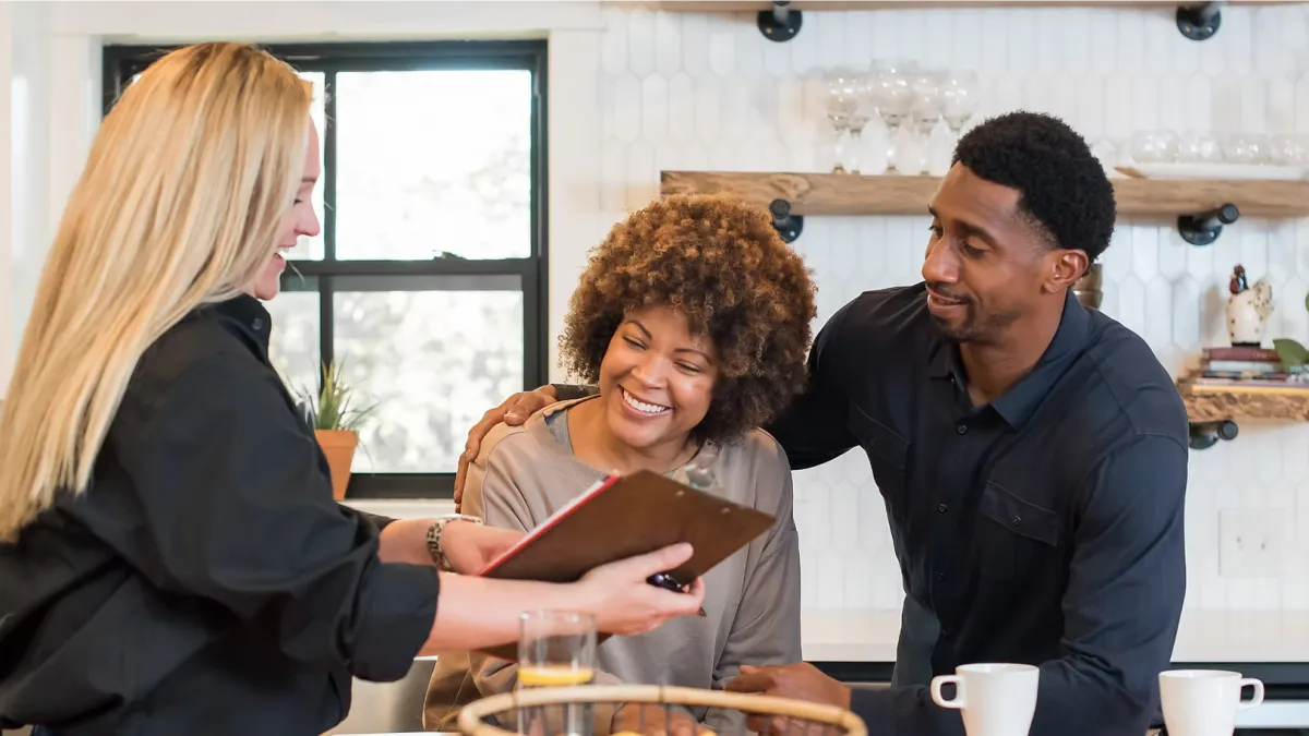 Couple smiling and reviewing a menu with a server in a modern kitchen setting with drinks on the counter