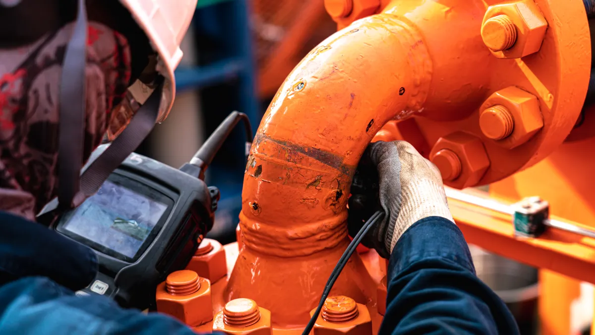 Worker inspecting large orange industrial pipe with handheld device wearing protective gear and gloves