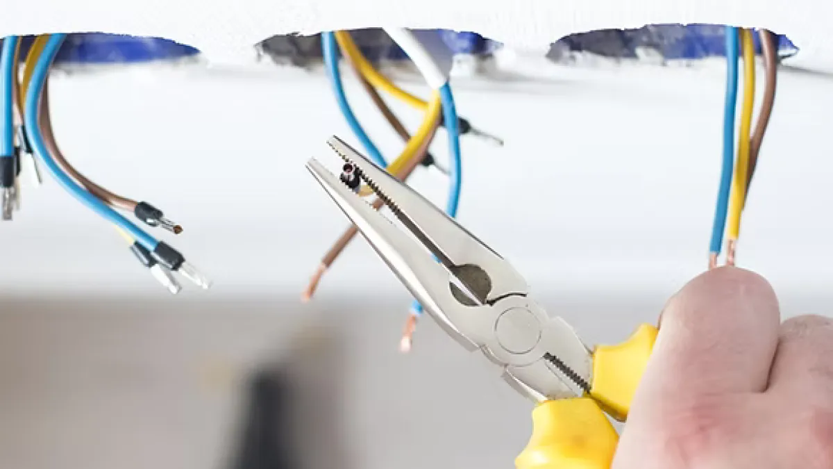Hand using yellow-handled pliers to work on exposed blue, yellow, and brown electrical wires in a wall.