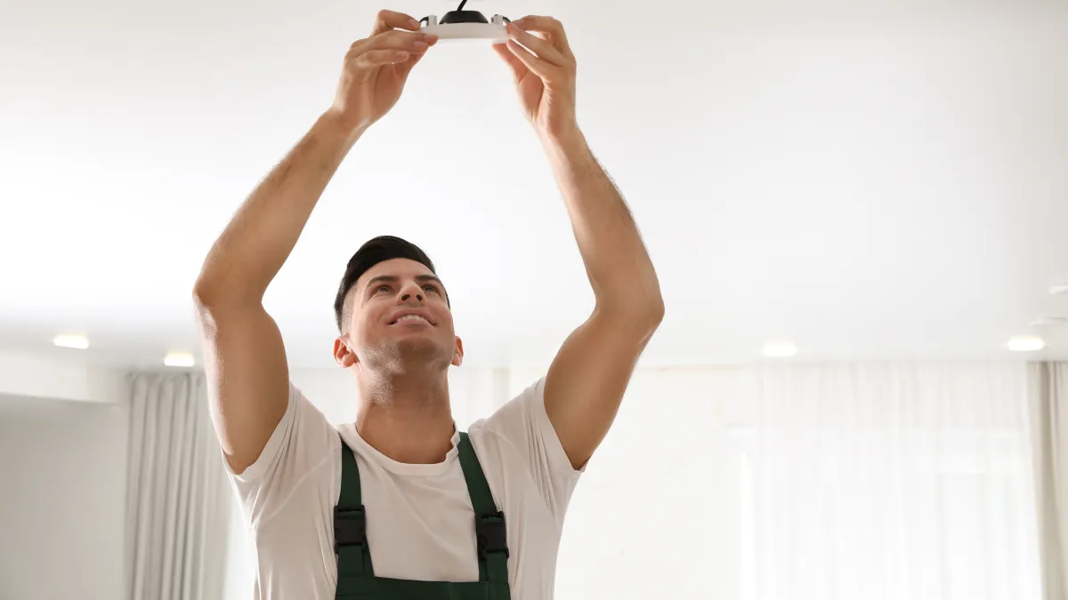 Technician installing or replacing a smoke detector on a white ceiling in a bright room.
