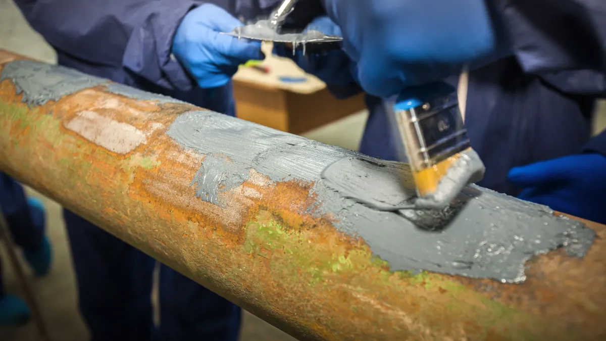 Workers applying gray anti-corrosion coating on a rusty metal pipe with brushes wearing blue gloves.