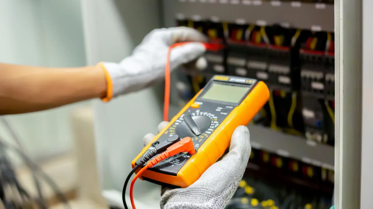 Electrician wearing gloves using a digital multimeter testing electrical panel wiring connections.