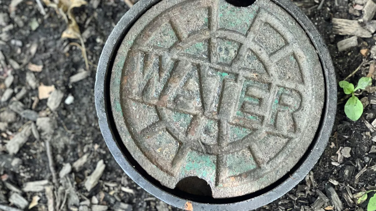 Rusty circular water valve cover embedded in soil with small green plants around.