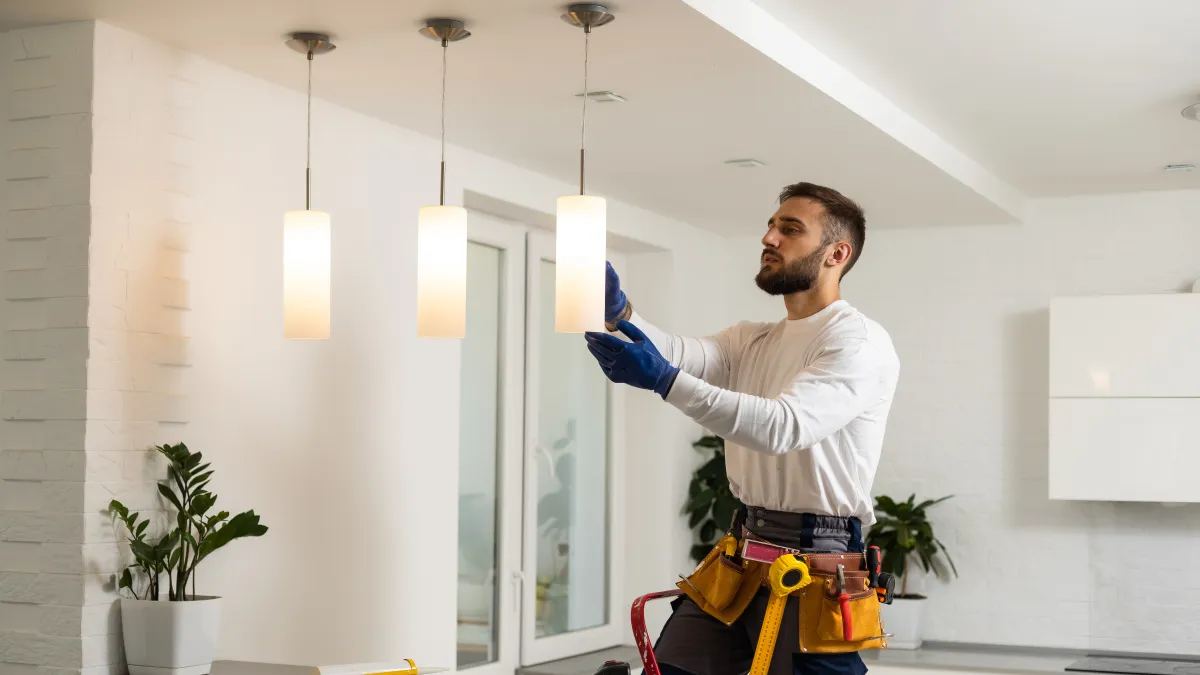 Electrician installing modern pendant lights in a bright, white kitchen interior with plants and tools on the counter.