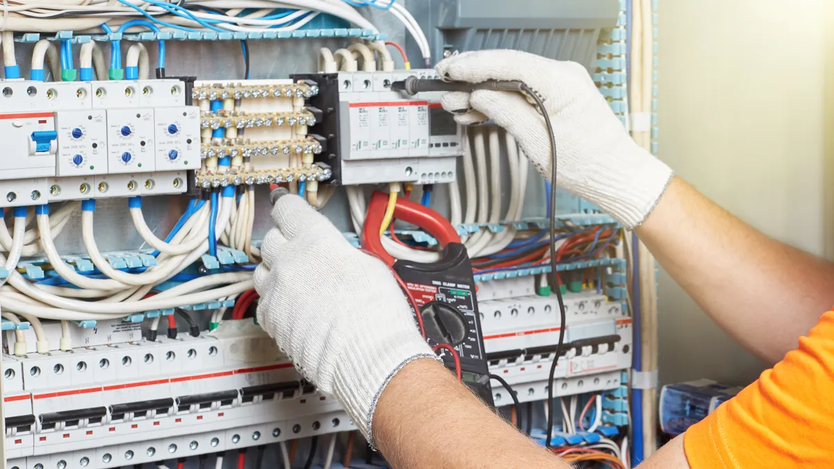 Electrician using multimeter to test circuit breakers and wiring inside electrical panel with gloves