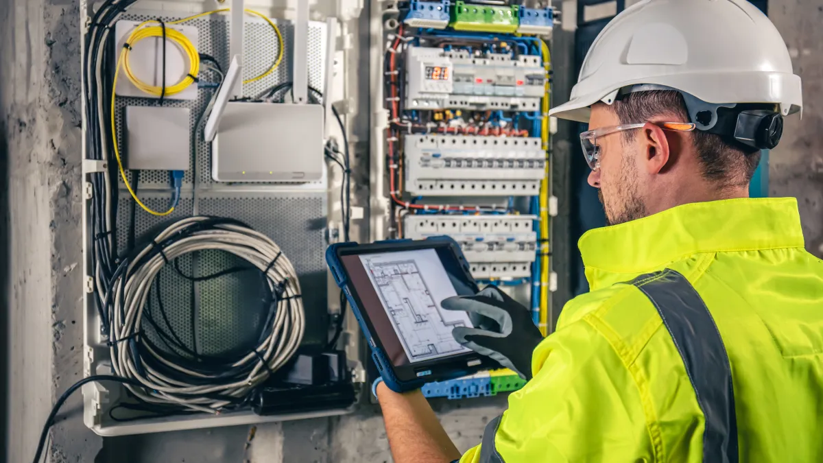 Electrician in safety gear inspecting wiring and circuit breakers using a tablet in a building under construction.