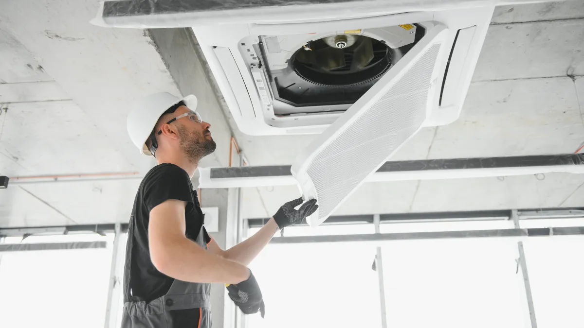 Technician in safety gear inspecting and removing ceiling air conditioning unit panel in industrial setting