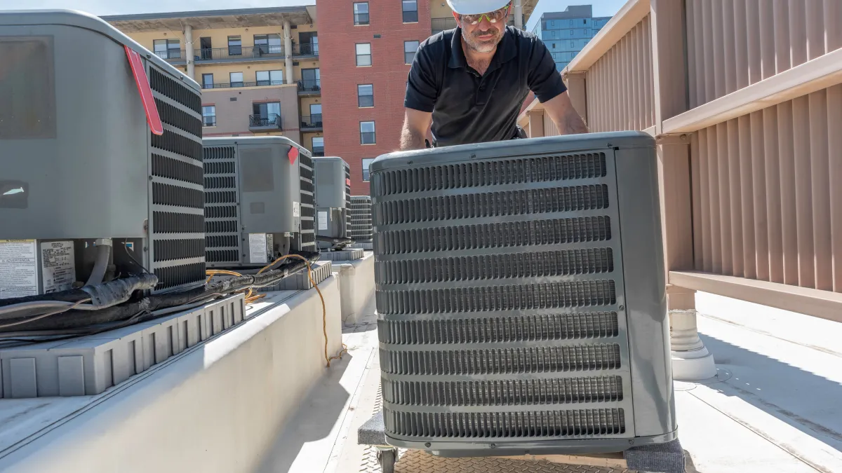 Technician in hard hat inspecting rooftop HVAC units on a sunny day near urban buildings.