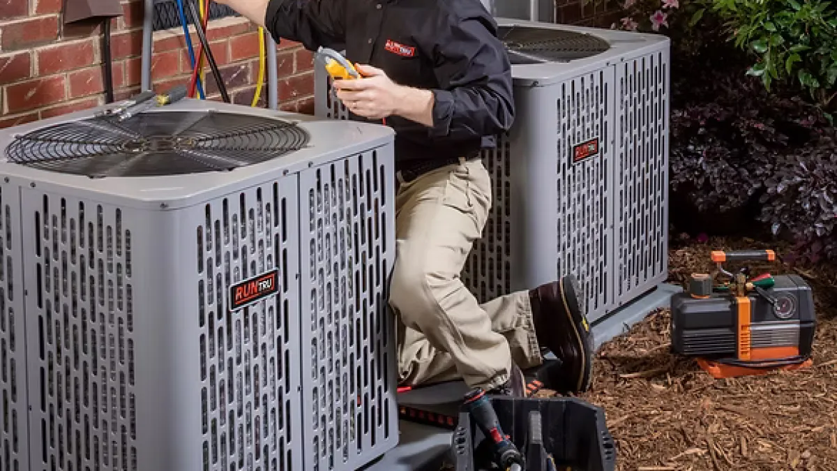 Technician in black shirt servicing two outdoor air conditioning units with tools and gauges near a house wall.