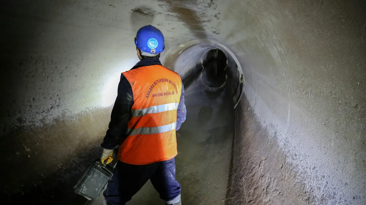 Worker wearing a hard hat and safety vest inspects a large underground drainage tunnel with flashlight.
