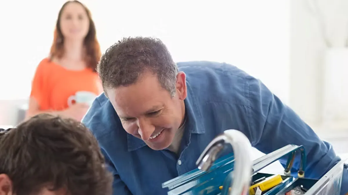 Two plumbers repairing a kitchen sink faucet with tools from a blue toolbox while a woman watches in the background