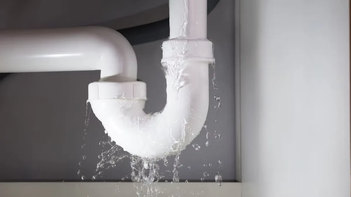 Leaking white PVC pipe under sink dripping water, showing plumbing issue and water damage risk.