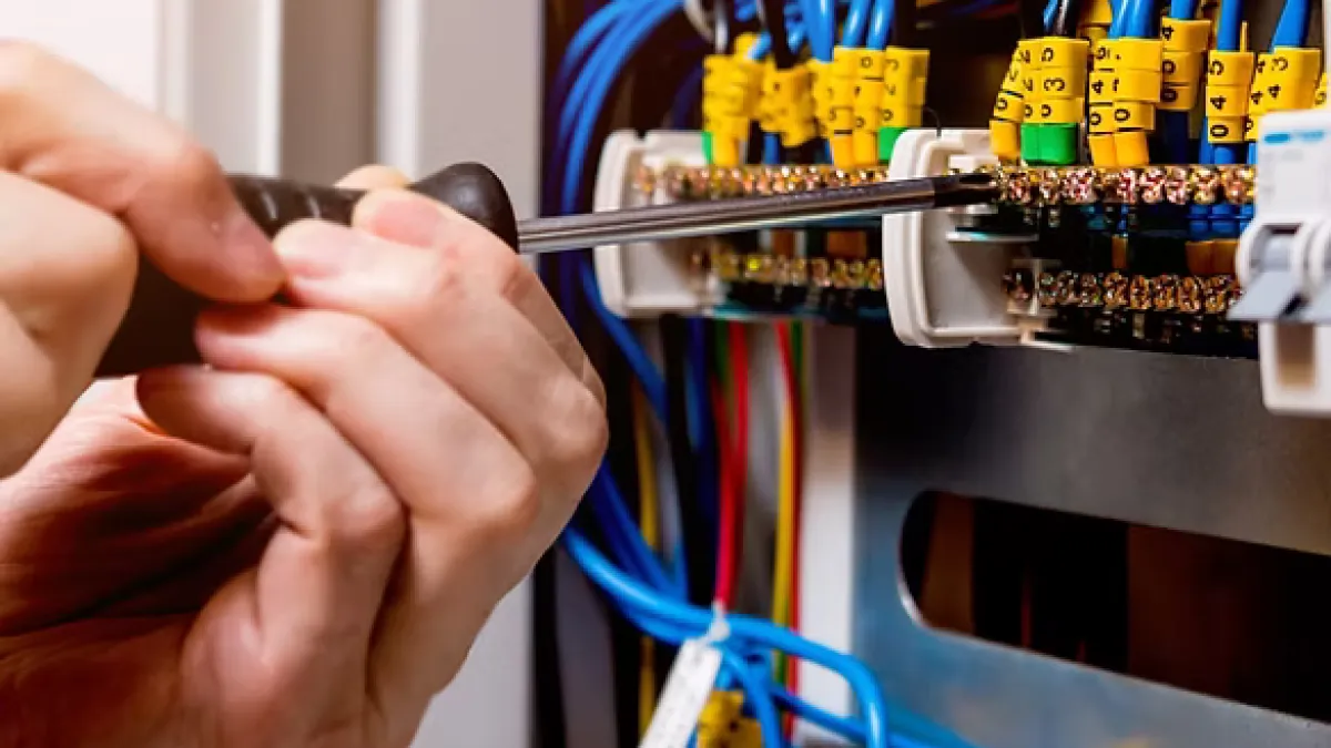 Electrician tightening screws on wiring panel with blue cables and yellow tags using a screwdriver indoors.