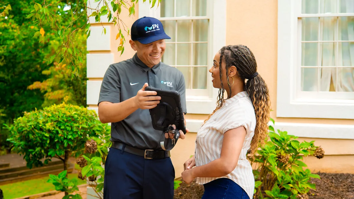 Technician in uniform showing a tablet to a woman outside a house with greenery and windows in the background