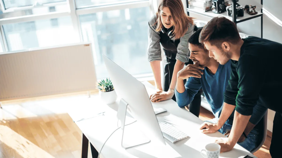 Three colleagues collaborating and discussing work on a desktop computer in a bright modern office.
