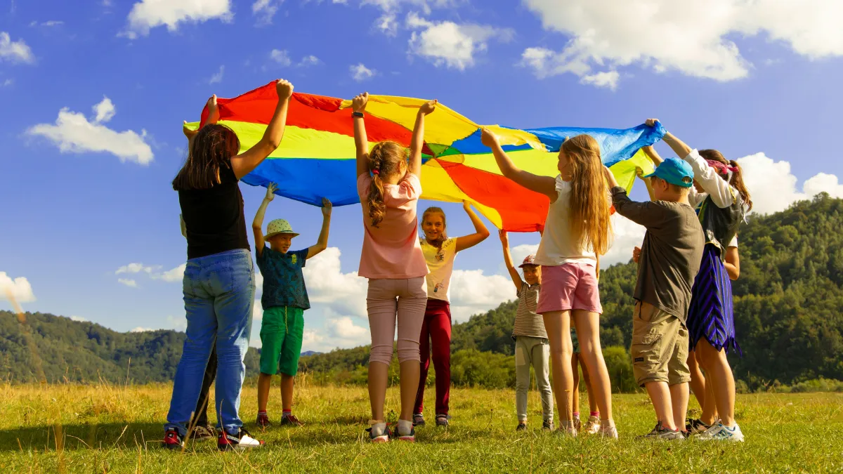 Children playing with a colorful parachute outdoors on a sunny day in a grassy field with hills in the background.