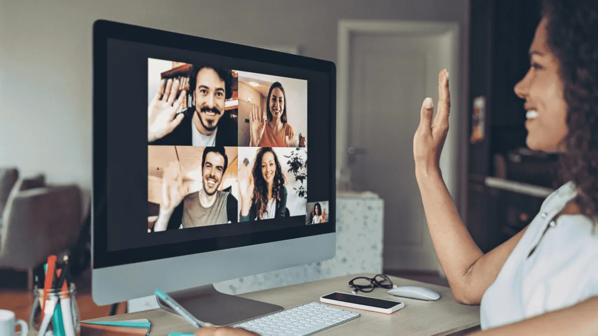 Woman waving and smiling during a video call with four friends on a desktop computer in a cozy home office.