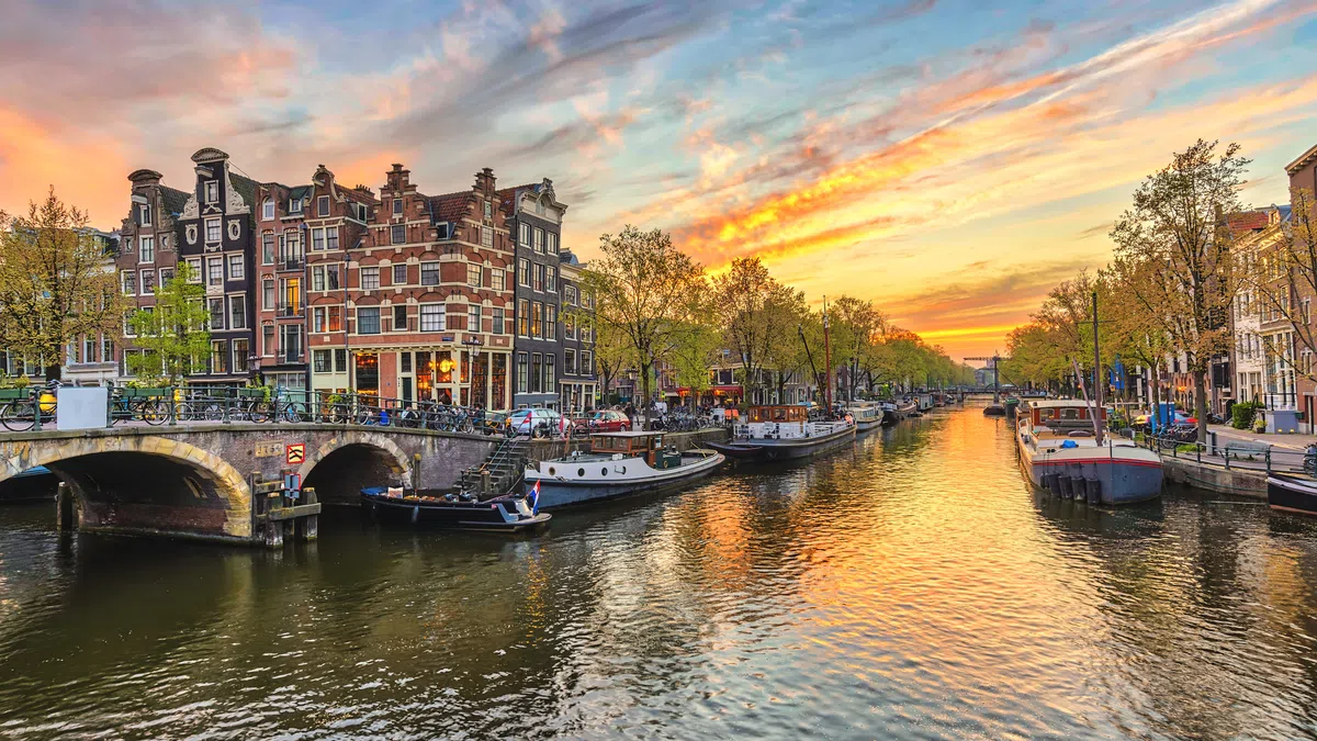 Sunset over Amsterdam canal with historic buildings, boats docked along tree-lined waterways and a stone bridge.