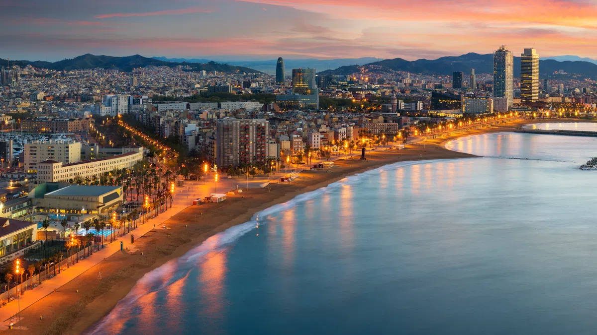 Panoramic view of Barcelona coastline at sunset with city skyline, beach, and calm sea under colorful sky