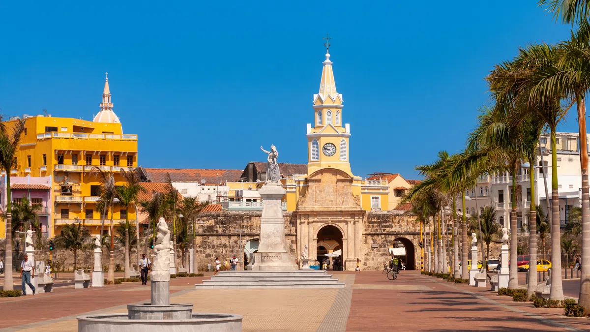 Historic yellow clock tower and statue in a sunny plaza lined with palm trees under a clear blue sky.