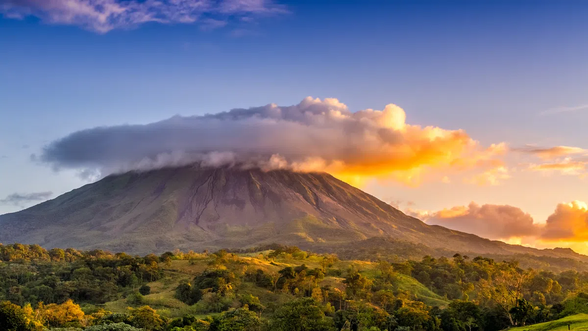 Sunset view of a mountain with clouds covering the peak and lush green forested hills in the foreground