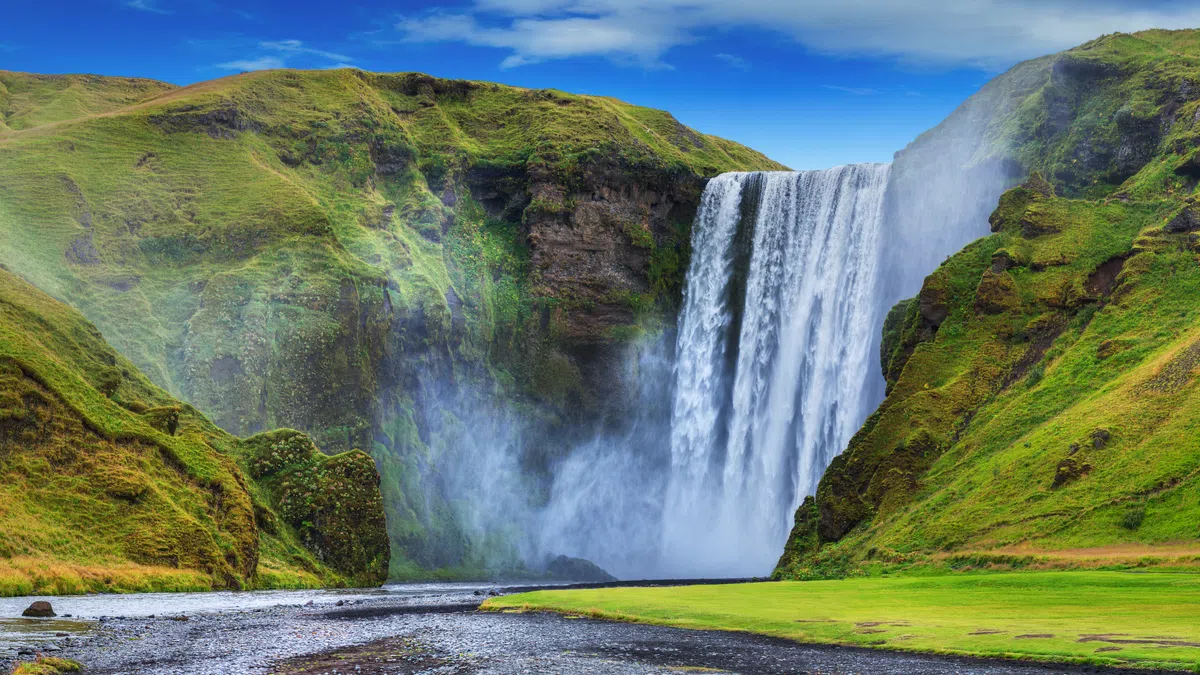 Wide view of a majestic waterfall cascading between lush green cliffs under a bright blue sky.